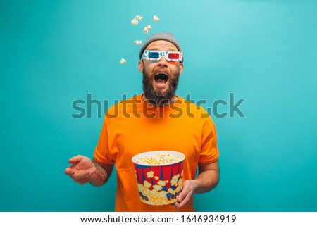 Handsome emotional young guy in orange t-shirt eat popcorn wearing 3d glasses watch film, isolated on blue background.