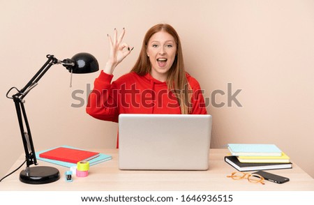 Young student woman in a workplace with a laptop surprised and showing ok sign