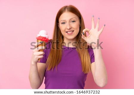 Young redhead woman with a cornet ice cream over isolated pink background showing ok sign with fingers