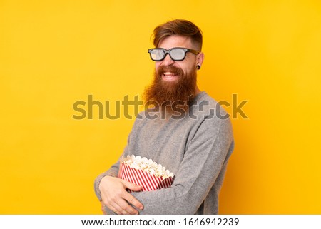 Redhead man with long beard over isolated yellow background with 3d glasses and holding a big bucket of popcorns