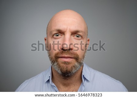 Keep calm and pretend nothing happened. Mature male with beard standing with no emotions over gray background, staring at camera without interest.