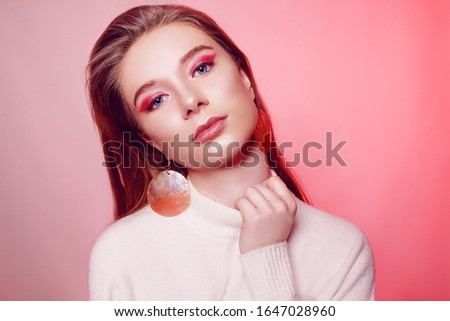 Incredibly beautiful blonde on a pink background. Portrait of baby face model. Girl with licked hair and big earrings. Jewelry. Girl in a white sweater.