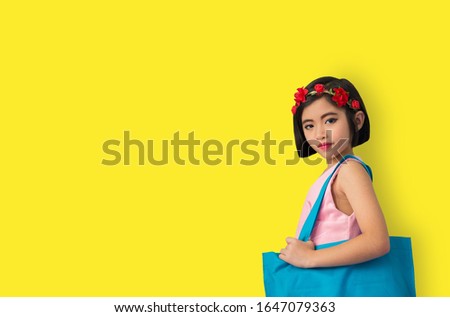 The girl in a pink dress is carrying a blue bag on his shoulder and a red flower on his head. Yellow studio background