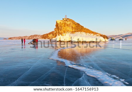 February is a good time to travel on the ice of the frozen Lake Baikal. A group of tourists photographs the beautiful rock Kobyliya Golova at sunset. Two all-terrain vehicles Hivus stand on the side 