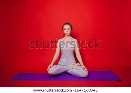 Athletic yoga, beautiful young girl practicing padmasana lotus position. Woman in black sportswear on a simple red background, standing on a purple carpet