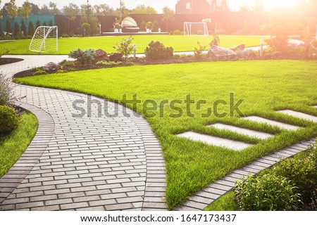 Landscaping of the garden. A tile path between green grass and a lawn with flowers in the sun. Soccer field in the background with copy space. Royalty-Free Stock Photo #1647173437