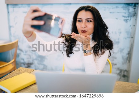 Pretty female millennial sending air kiss while making online web conference for attracting social followers, cute Caucasian hipster girl using cellphone gadget for clicking selfie pictures