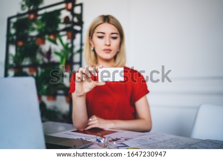 Elegant smart woman attentively studying business card in hand and reading information while working with documents at table in office 