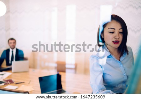 Through glass pensive Asian adult businesswoman explaining opinion for blurred coworker in formal clothes while standing and writing on marker board during negotiation in modern conference room