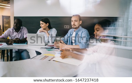 Smiling colleagues sitting at table loaded with laptop notebooks opposite of green board in office space seen from window 
