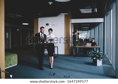Man in formal clothes speaking to beautiful female in glasses with tablet in hands while standing in office building hallway