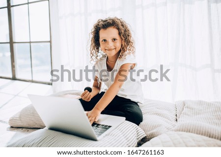 Cheerful adorable curly haired child in casual clothes surfing on laptop eating cookies sitting on pillows in light living room in sunny day