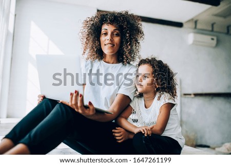 Content ethnic woman with curly hair using laptop at white apartment and looking at camera while little daughter sitting beside and watching at screen with interest