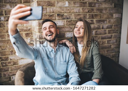 Confident smiling modern woman and man in denim shirt sitting together on couch against brick wall and capturing selfie in cafe 
