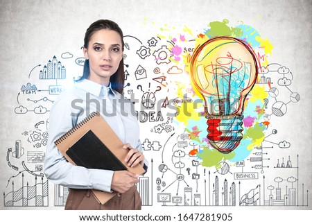 Serious young woman with dark hair and notebooks standing near concrete wall with colorful business idea sketch. Concept of creativity and startup
