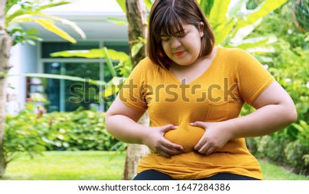 Overweight young woman pinches fat on her belly large size while sitting in the garden at home. Upset female suffering from extra weight. Obesity unhealthily concept. Royalty-Free Stock Photo #1647284386