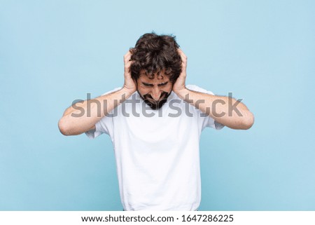 young crazy bearded man feeling stressed and frustrated, raising hands to head, feeling tired, unhappy and with migraine against flat wall
