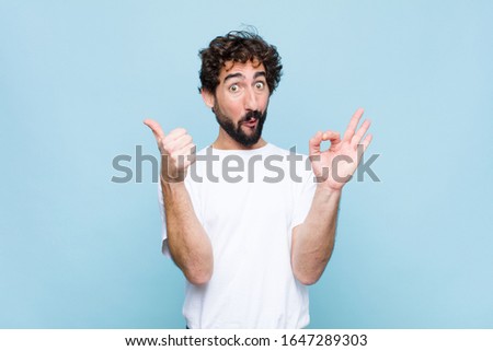 young crazy bearded man feeling happy, amazed, satisfied and surprised, showing okay and thumbs up gestures, smiling against flat wall