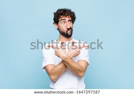 young crazy bearded man looking puzzled and confused, insecure and pointing in opposite directions with doubts against flat wall