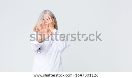 middle age doctor woman covering face with hand and putting other hand up front to stop camera, refusing photos or pictures