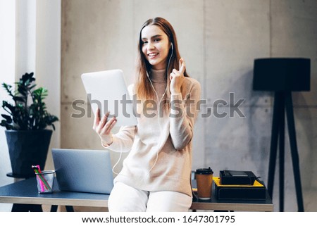Female manager sitting on office desk and performing corporate tasks using wireless connection on digital gadgets. Girl listening to music, watching video in earphones and looking at screen of tablet.