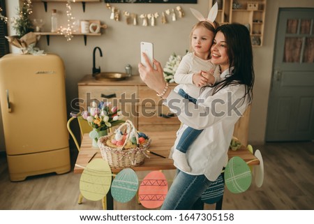 Happy mother with daughter in her arms making selfie on smartphone on wooden kitchen background and easter eggs and tulips