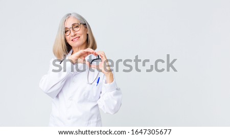 middle age doctor woman smiling and feeling happy, cute, romantic and in love, making heart shape with both hands