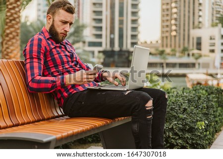 A handsome guy with a beard is sitting on the bench. He holds a laptop in his lap and uses a cell phone
