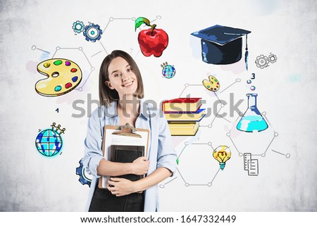 Smiling teen girl in smart casual clothes standing with books near concrete wall with colorful education sketch drawn on it. Concept of career and education