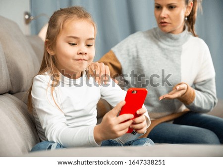 Small daughter looking at phone, mother on background  at home  interior