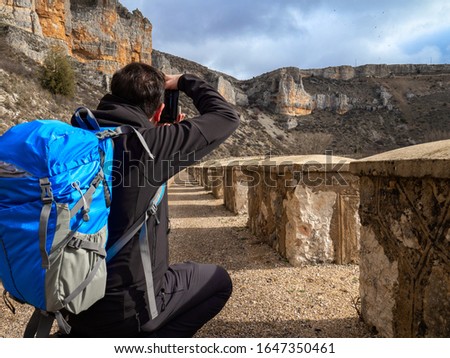 Photographer with a blue bag crouching in the middle of a sandy path taking a picture in the middle of the mountain surrounded by rock formations in Maderuelo in Spain