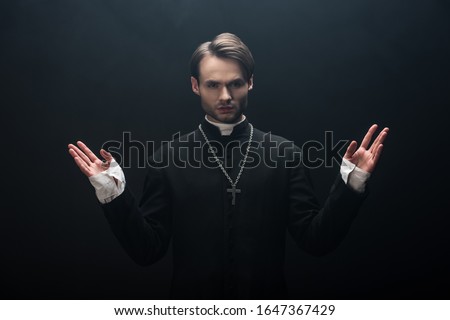 young confident catholic priest looking at camera while standing with open arms isolated on black