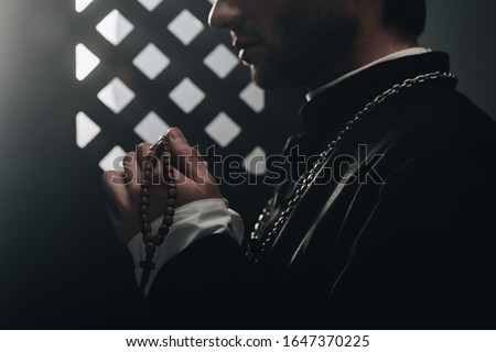 partial view of catholic priest holding wooden rosary beads near confessional grille in dark with rays of light Royalty-Free Stock Photo #1647370225