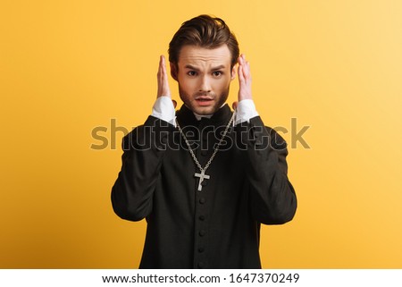 shocked catholic priest holding hands near head and looking at camera isolated on yellow