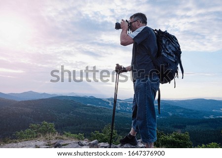 Tourist hiker photographer with backpack and hiking poles takes pictures on the top of rocky mountain, enjoying view of valley covered with white clouds stretching to horizon.