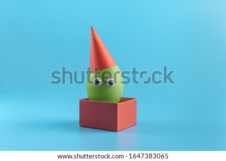 One green egg with eyes in a red festive cap in a square red box on a blue background. Minimal Happy Easter concept decoration. Copy space for text mockup. Close up photography. 