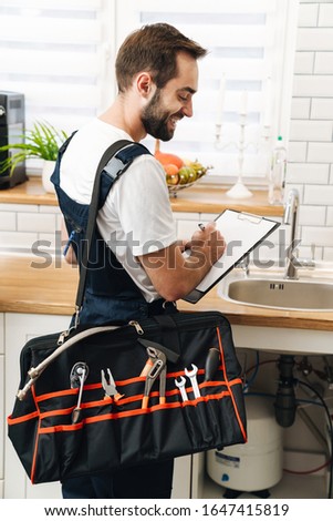Image of young man plumber work in uniform indoors holding bag with equipment and clipboard.