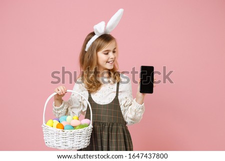 Little blonde kid girl 11-12 years old in spring dress bunny ears hold in hand cell phone wicker basket colorful eggs isolated on pastel pink background Childhood lifestyle Happy Easter concept