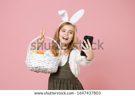 Little blonde kid girl 11-12 years old in dress bunny rabbit ears hold in hand cell phone wicker basket colorful eggs isolated on pastel pink background Childhood lifestyle Happy Easter concept