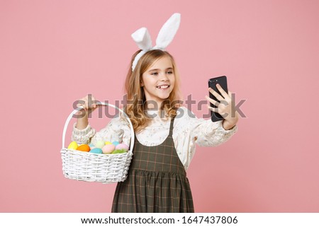 Little pretty blonde kid girl 11-12 years old in spring dress, bunny rabbit ears hold in hand wicker basket colorful eggs isolated on pastel pink background. Childhood lifestyle Happy Easter concept