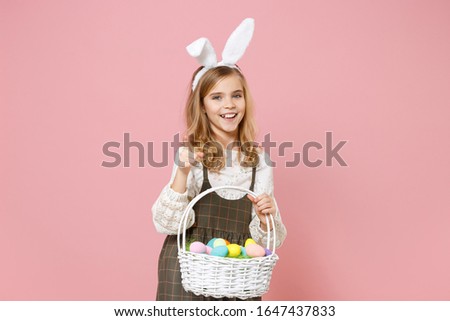 Little pretty blonde kid girl 11-12 years old in spring dress, bunny rabbit ears hold in hand wicker basket colorful eggs isolated on pastel pink background. Childhood lifestyle Happy Easter concept