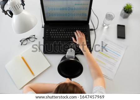 technology, mass media and people concept - woman with microphone and laptop computer reading text from papers and recording podcast at studio