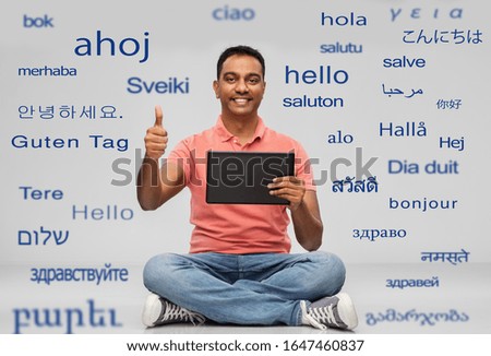 technology, communication and people concept - happy indian man with tablet computer sitting on floor over greeting words in different foreign languages on grey background