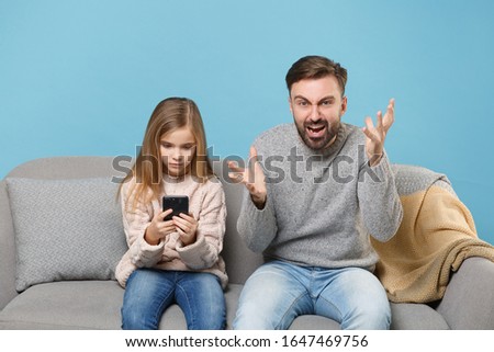 Angry bearded man in knitted sweater with child baby girl. Father little daughter isolated on pastel blue background. Love family parenthood childhood concept. Sitting on couch, hold mobile phone