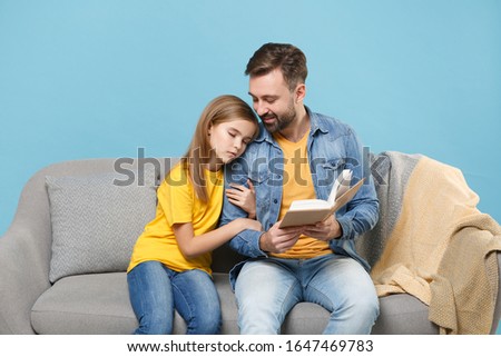 Smiling bearded man in casual clothes with cute child baby girl. Father little daughter isolated on pastel blue background. Love family parenthood childhood concept. Sit on couch reading book hugging