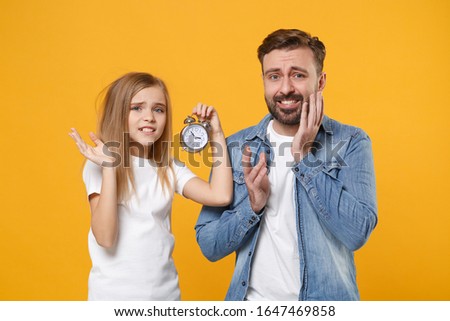 Concerned bearded man in casual clothes with child baby girl. Father little kid daughter isolated on yellow background. Love family parenthood childhood concept. Hold alarm clock look at each other