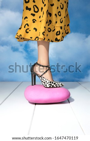 Smart girl standing on a pink stone maintaining her full balance while wearing mustard colored cheetah print frock and leopard design mules in pencil heels. Background is blue cloud & surface is white