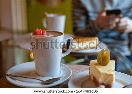 Close up of white cup of coffee latte decorated with a heart and flower, cake with bite taken. Man holding smart phone on background. Stock photo.