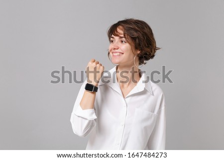 Smiling young business woman in white shirt posing isolated on grey background in studio. Achievement career wealth business concept. Mock up copy space. Wearing smart watch on hand, looking aside