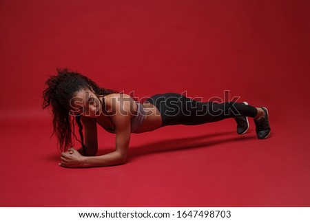 Strong attractive young african american sports fitness woman in sportswear working out isolated on red background studio portrait. Sport exercises healthy lifestyle concept. Doing plank exercises